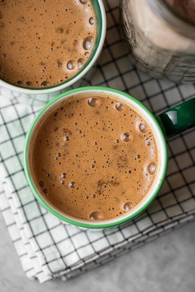 Xícara de cappuccino caseiro com chocolate, vista de cima, mostrando a textura cremosa e espumosa da bebida quente sobre uma toalha xadrez.