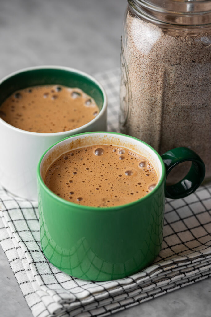 Duas xícaras de cappuccino caseiro com chocolate ao lado de um pote de vidro com a mistura pronta, sobre uma toalha branca com listras pretas.