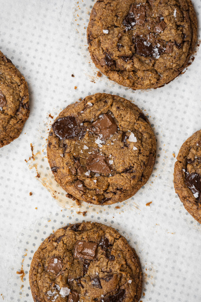 Cookies chips com manteiga tostada e pedaços de chocolate, dispostos sobre papel manteiga perfurado. As bordas estão douradas e o centro levemente derretido, finalizados com flor de sal.