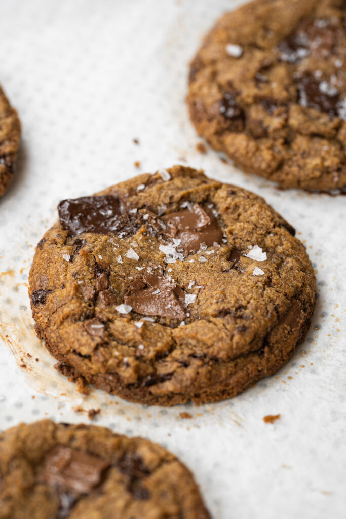 Close de um cookie chips com manteiga tostada, mostrando textura crocante nas bordas e pedaços de chocolate derretido no topo, com flor de sal salpicada por cima.