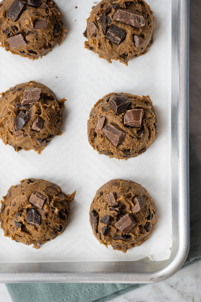 Bolinhas de massa de cookie chips com manteiga tostada e pedaços de chocolate dispostas em uma assadeira forrada com papel manteiga, prontas para serem assadas.