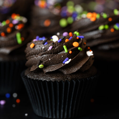Cupcake Black Velvet decorado com cobertura cremosa e confeitos de Halloween nas cores laranja, verde e roxo.