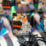 Docinhos de fudge de cookies and cream com recheio laranja e confeitos coloridos, decorados com aranha plástica e papel iridescente para o Halloween.