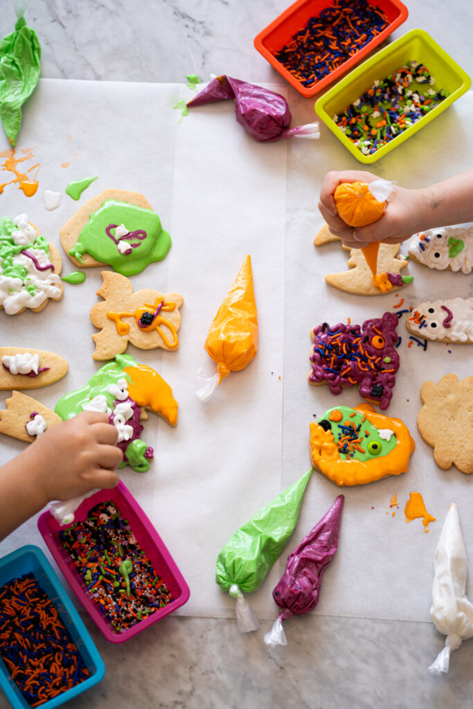 Crianças decorando biscoitos de Halloween com glacês coloridos e confeitos variados sobre papel manteiga.