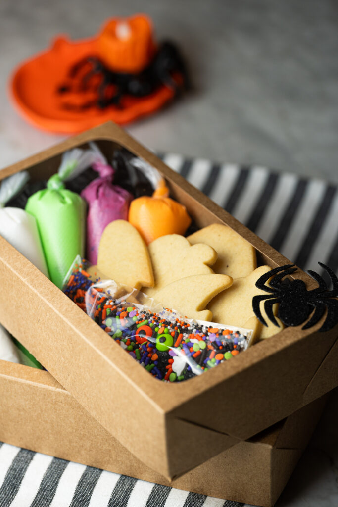 Halloween Cookie Box com biscoitos amanteigados, saquinhos de glacê colorido e confeitos temáticos dentro de uma caixa kraft decorada.