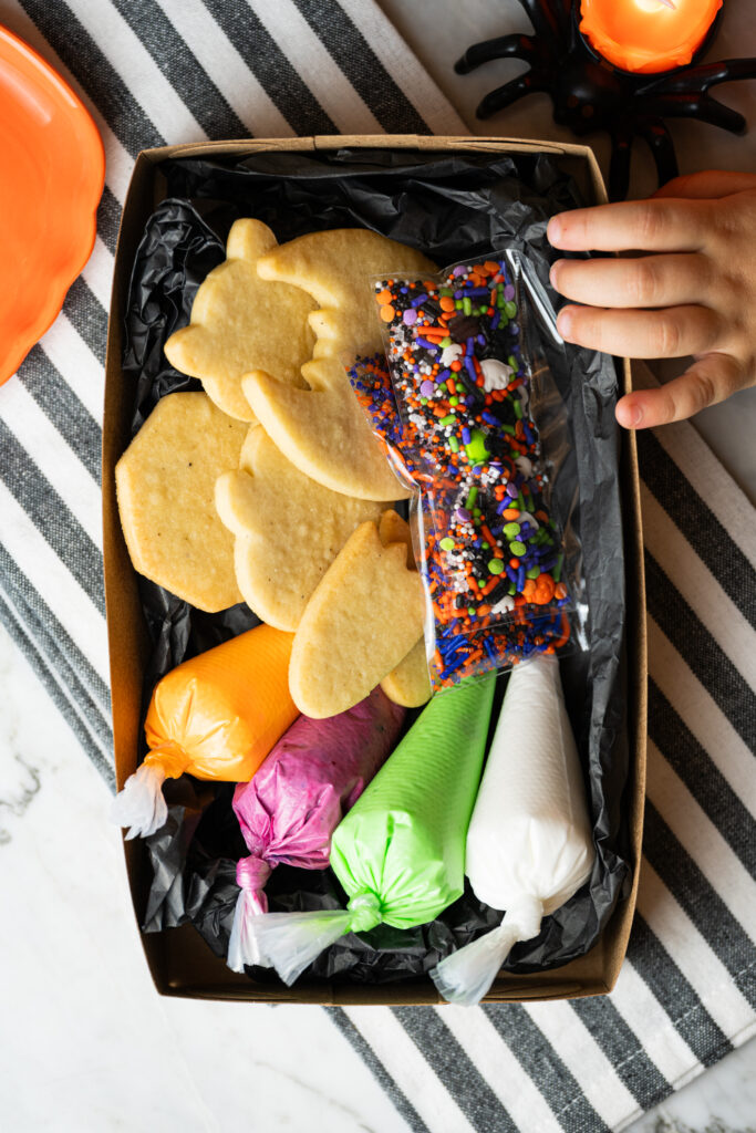 Vista de cima da Halloween Cookie Box aberta, mostrando biscoitos prontos para decorar, confeitos e glacês coloridos embalados.