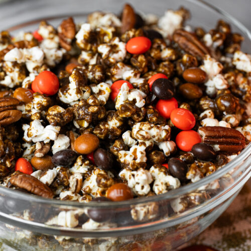 Tigela de vidro cheia de Pipoca de Pumpkin Spice caramelizada com M&Ms e nozes pecan sobre toalha com bordado de abóbora, perfeita para o outono e o Halloween.