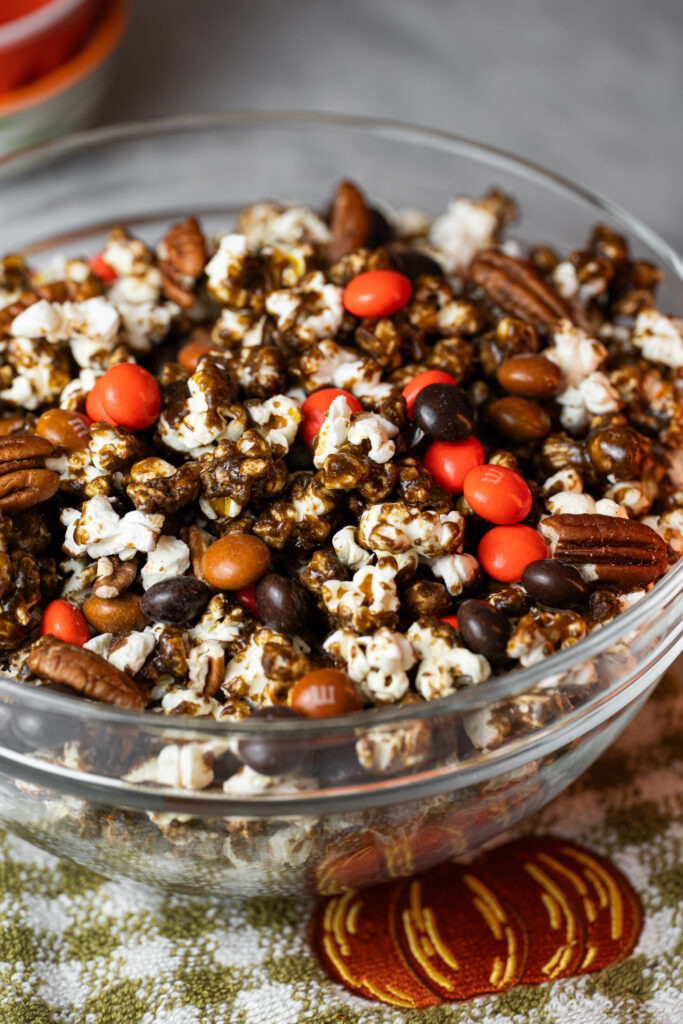 Tigela de vidro cheia de Pipoca de Pumpkin Spice caramelizada com M&Ms e nozes pecan sobre toalha com bordado de abóbora, perfeita para o outono e o Halloween.