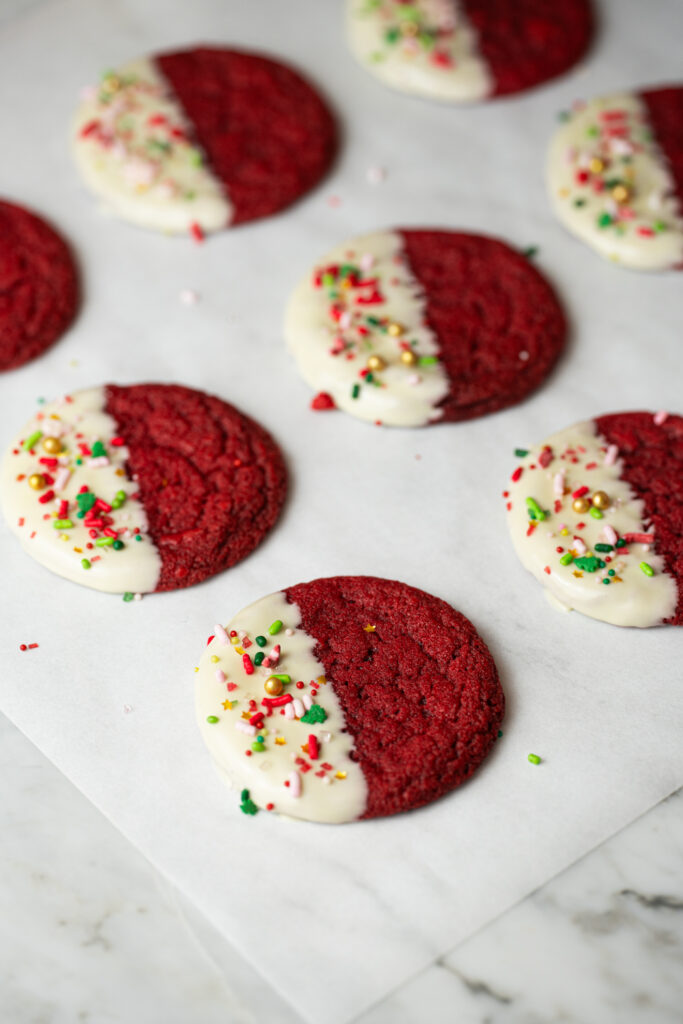 Cookies Red Velvet de Natal banhados em chocolate branco com confeitos coloridos, secando sobre papel manteiga.
