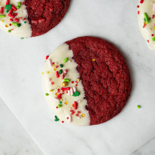 Cookie Red Velvet de Natal com metade banhada em chocolate branco e decorada com confeitos vermelhos, verdes e dourados.