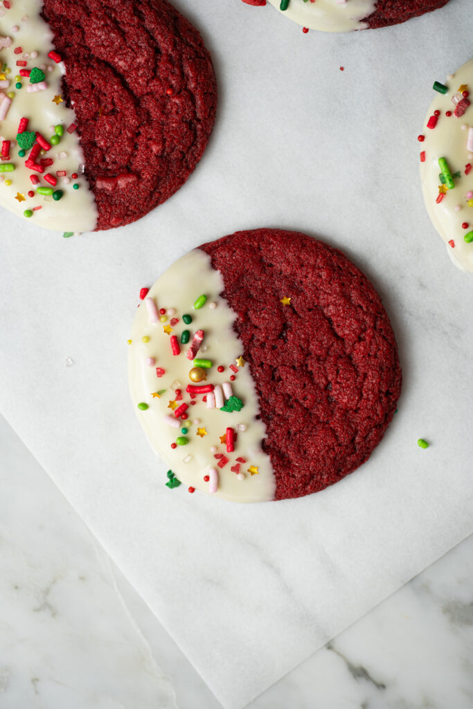 Cookie Red Velvet de Natal com metade banhada em chocolate branco e decorada com confeitos vermelhos, verdes e dourados.