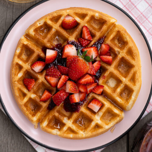 Waffle caseiro crocante e macio visto de cima, servido com geleia, morangos picados e mel sobre prato claro.