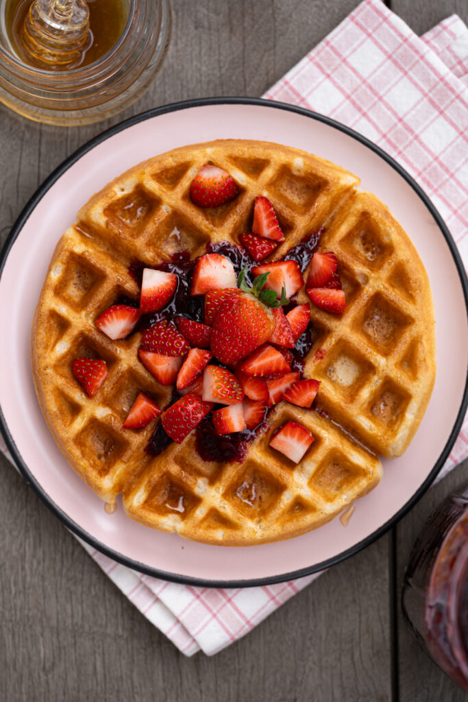 Waffle caseiro crocante e macio visto de cima, servido com geleia, morangos picados e mel sobre prato claro.